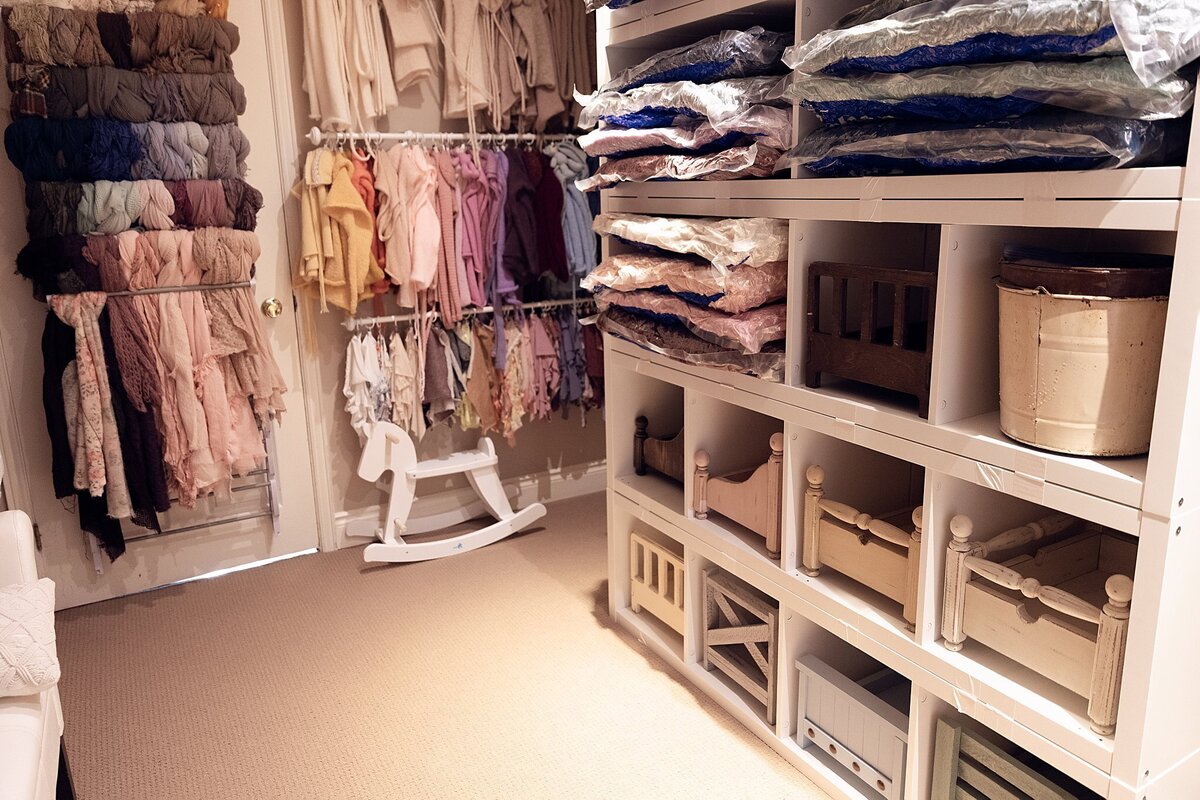 Organized shelving in a newborn photography studio displaying folded fabrics, outfits, wraps, and baskets, with a white rocker nearby.