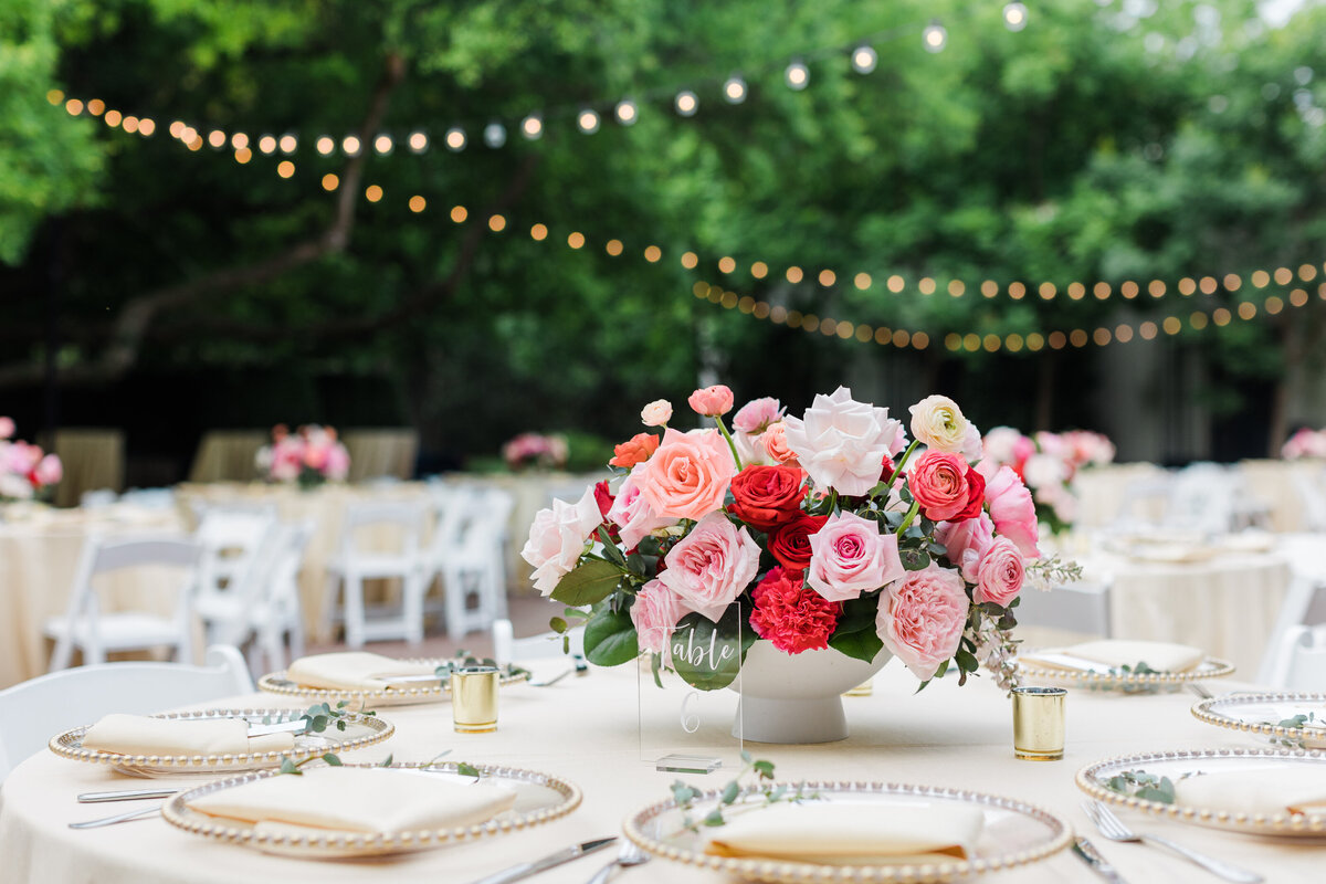 A detail shot of a reception table at a wedding reception at The Dallas Arboretum in Dallas, Texas. The large round table has a cream tablecloth, multiple fancy place settings, and a centerpiece made up of a large bouquet of pink, white, and red roses and multiple candles. Similar tables can be seen in the backyard while strings of cafe lights criss cross over head.