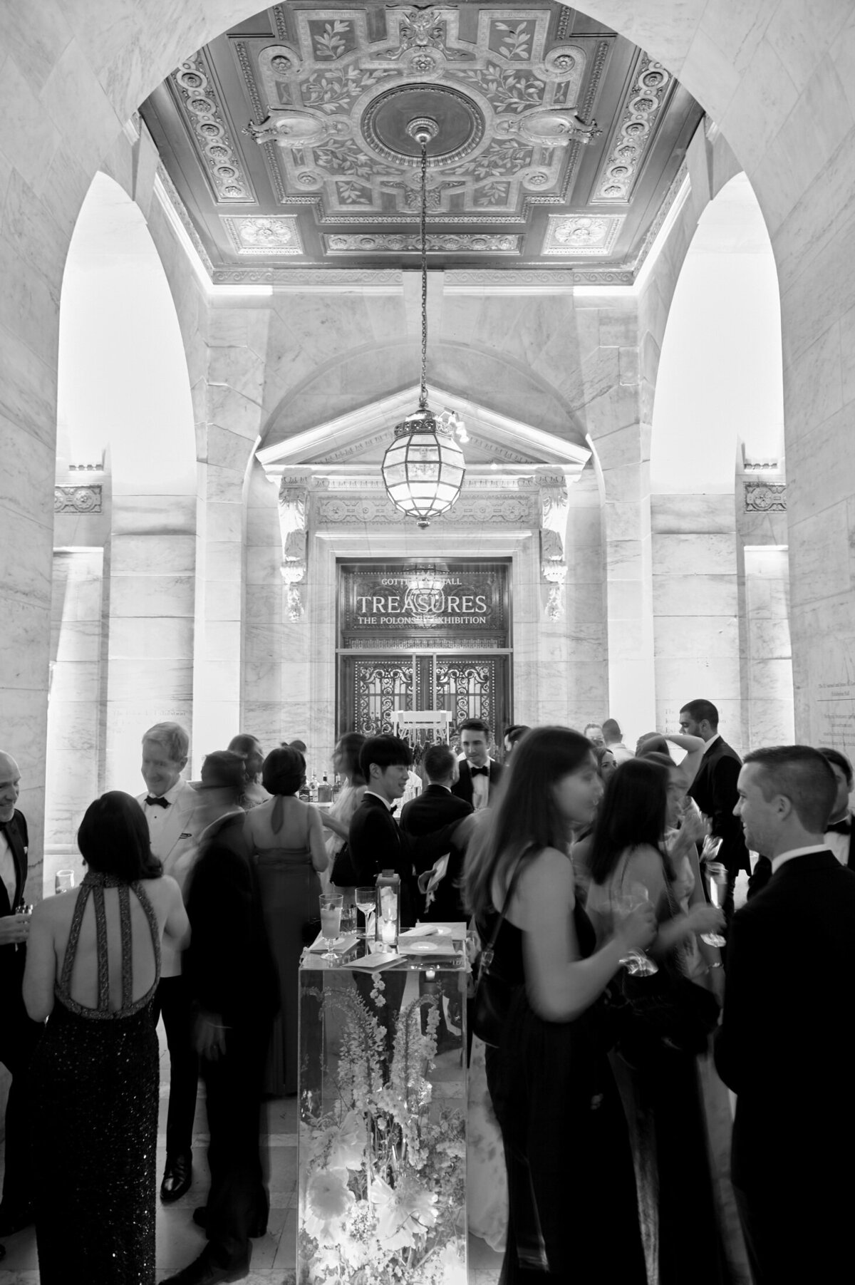 Black and white image of a formal event inside an ornate hall with high, elaborately decorated ceilings. People dressed in evening attire mingle and converse, some holding drinks. A lit display case in the center showcases decorative items. An arched entrance in the background.