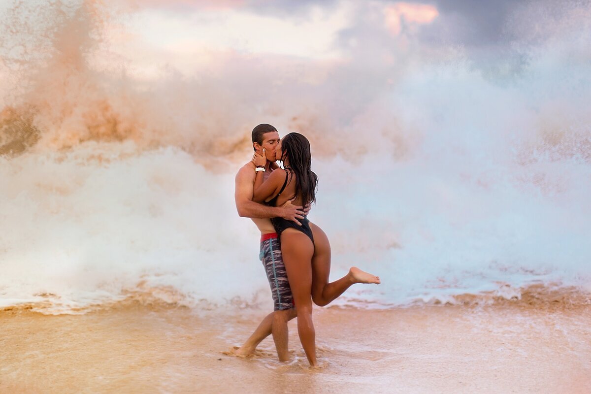 Couple kissing as a large shorebreak crashes behind them for their beach engagement session with Love + Water in Wailea