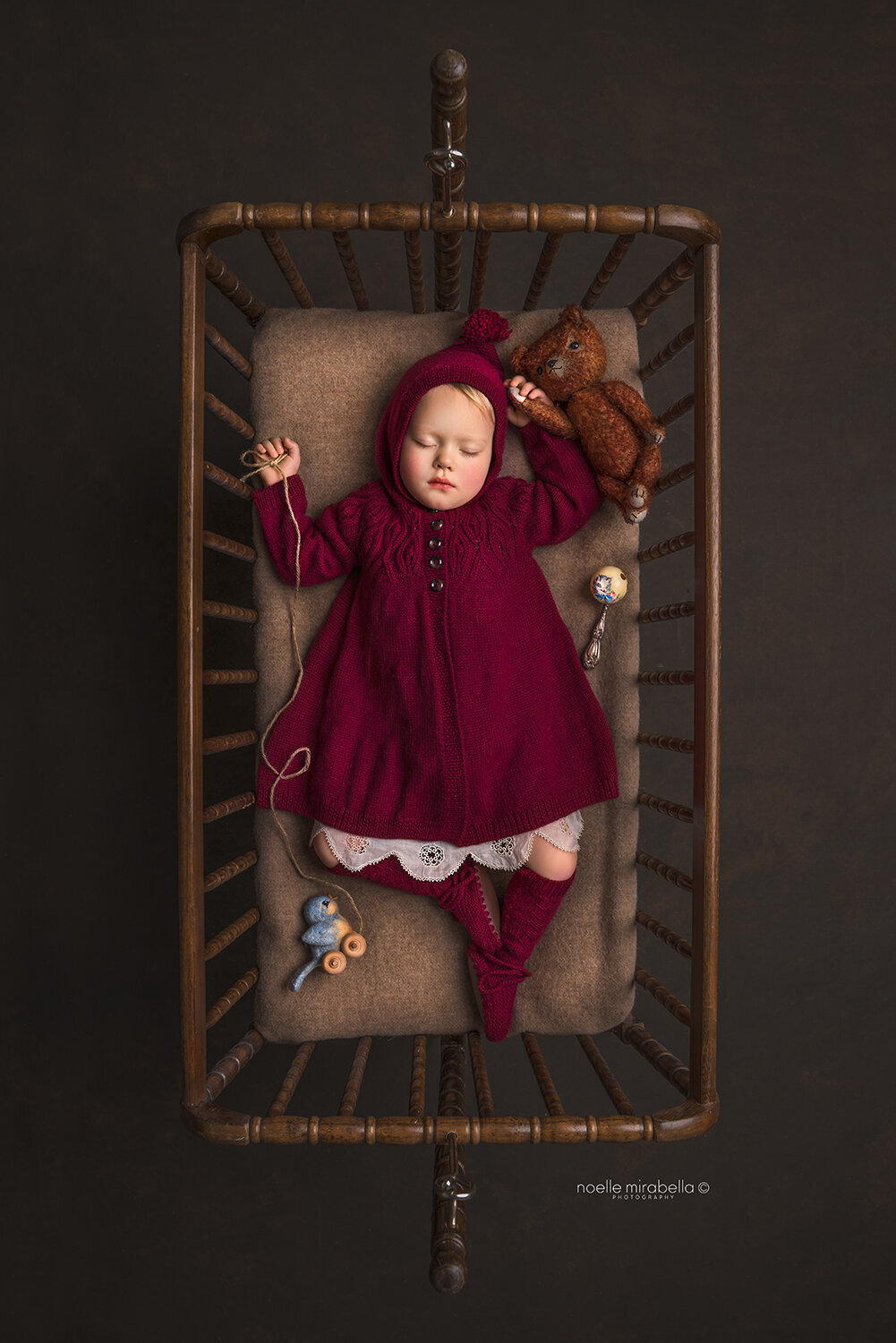 Toddler baby in red knits sleeping in vintage Jenny Lind cradle.