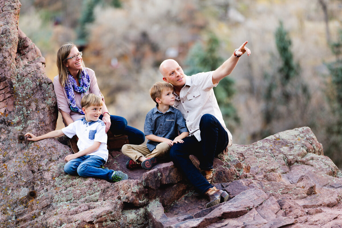 a dad points off into the distance while he, his two sons, and wife sit on pink rocks on a hill in boulder, colorado