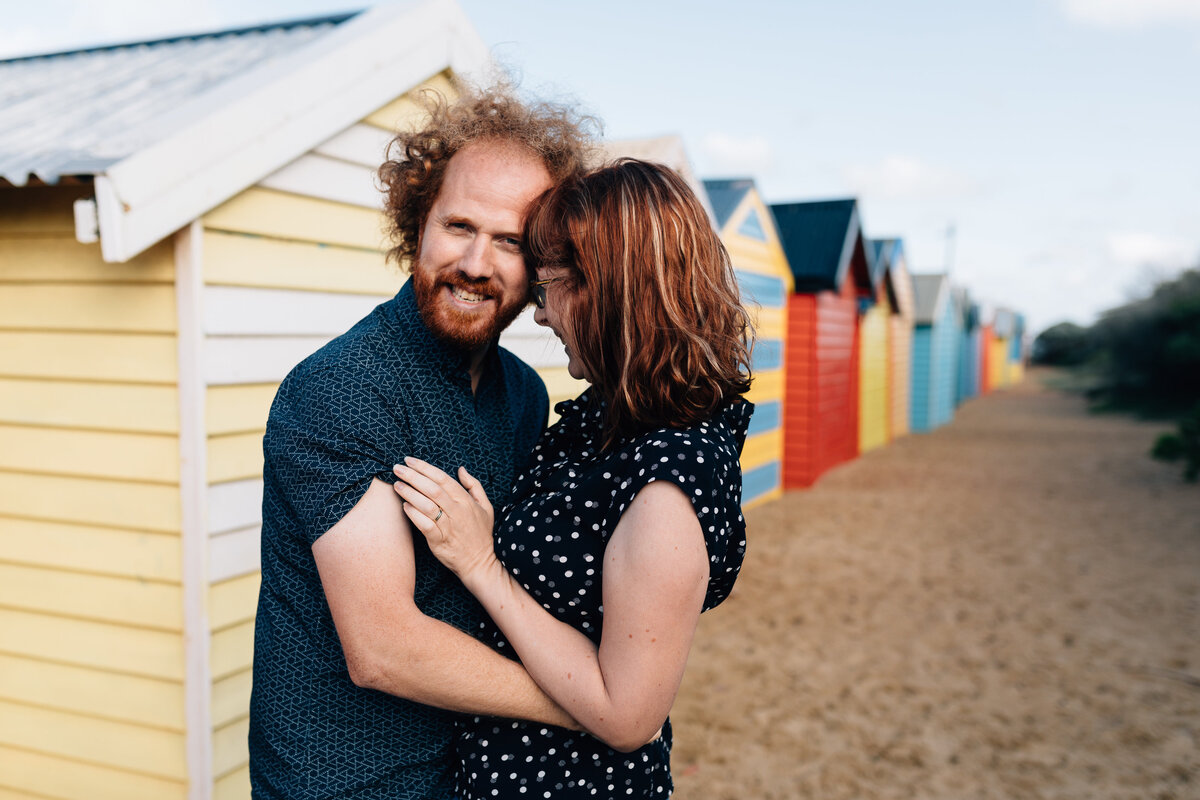 Couple hugging, man looking at camera, woman laughing into man at Brighton Beach Bathing Boxes. Couples photography Melbourne, And So I Don’t Forget Photography