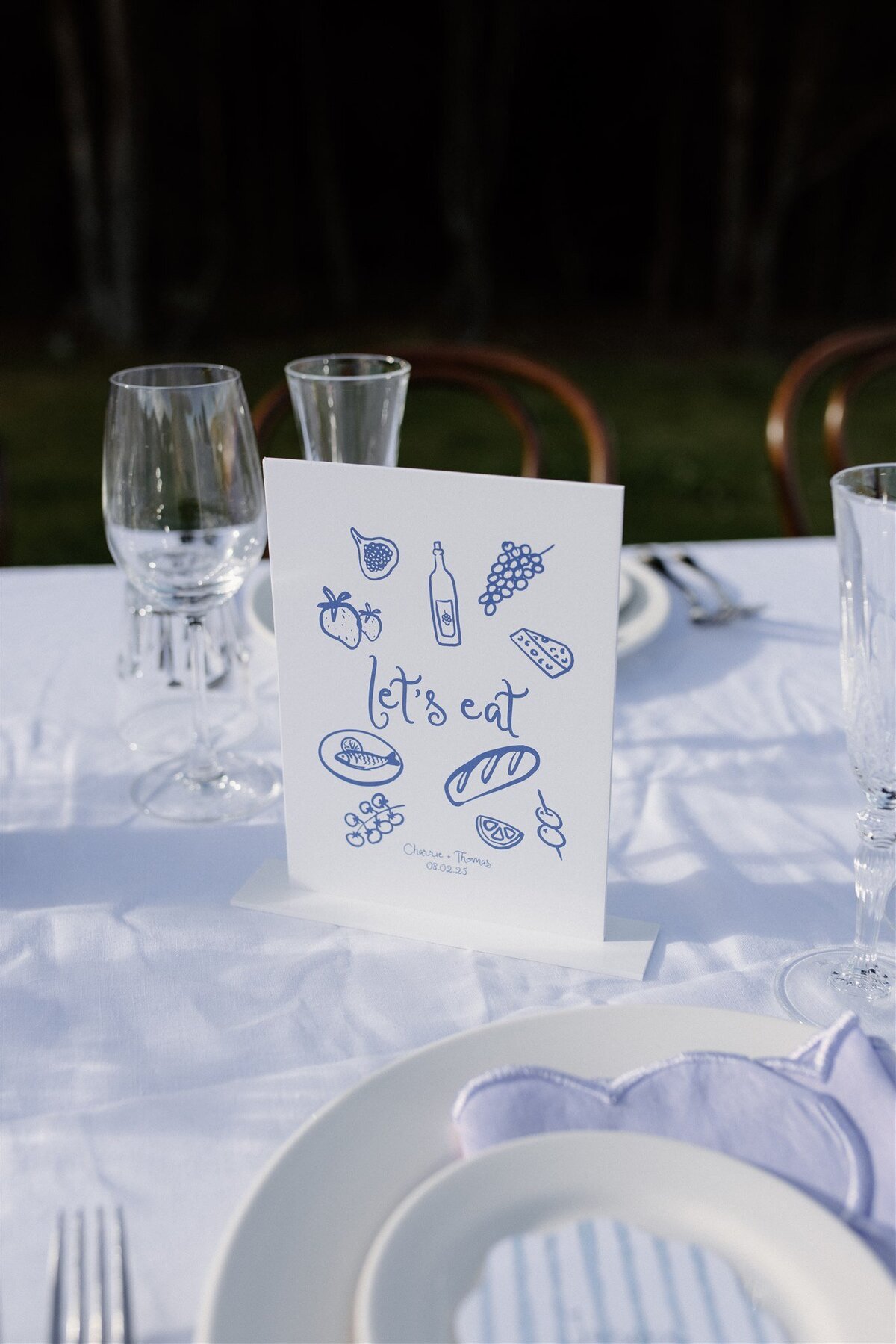 A playful and illustrated menu card reading “Let’s Eat” sits among elegant glassware, with figs, grapes, cheese, and bread drawings setting a European picnic tone. Personalised with the couple’s names and wedding date, it adds a relaxed, whimsical energy to the tablescape.