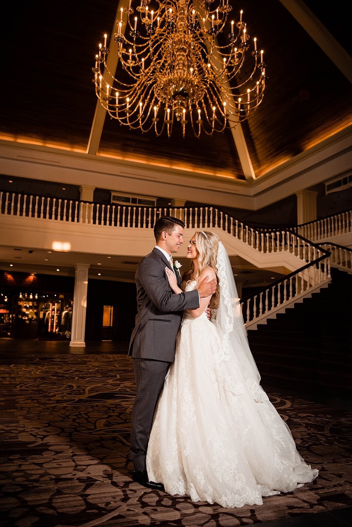Couple slow dancing under chandelier inside of the lobby with a staircase behind them