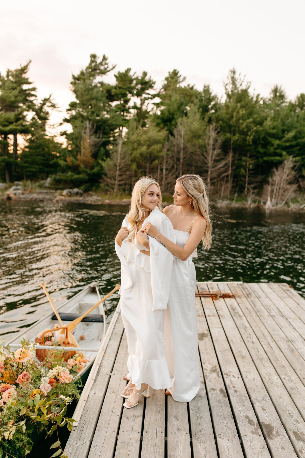 Two women in elegant white attire sharing a close embrace on a wooden dock over a tranquil lake surrounded by forest