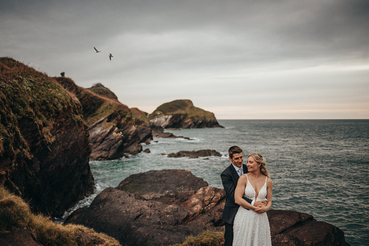 bride and groom embrace on the rocks by the ocean on their wedding day