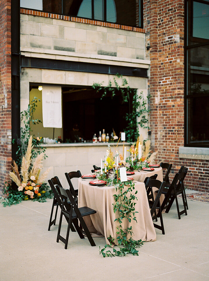 A curved table is set with ivory table cloth and green garland, red napkins and candlesticks outside.