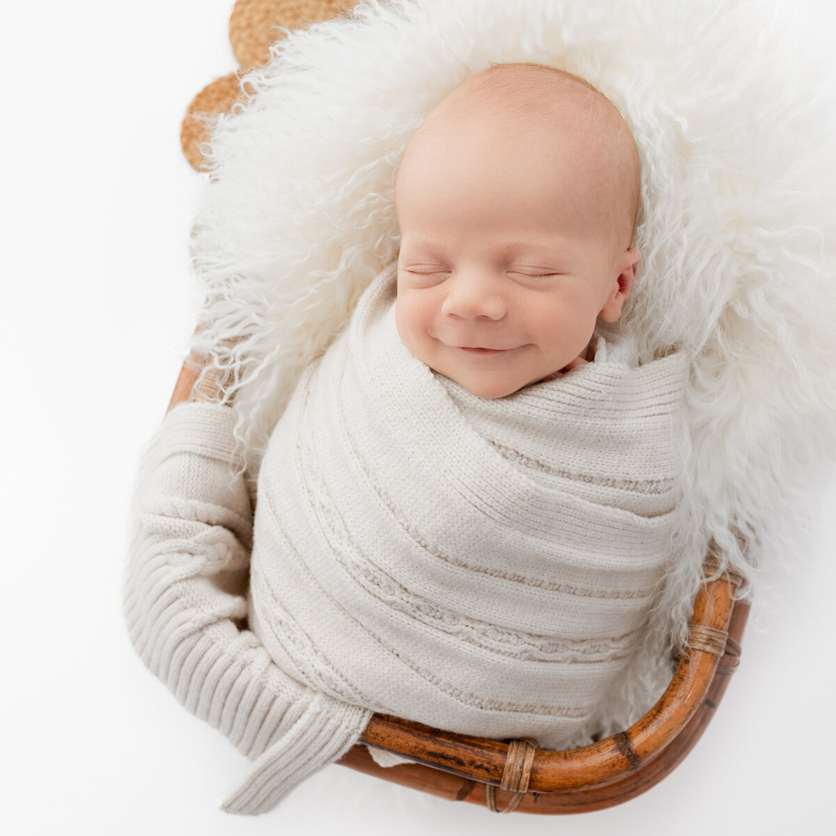 Image is of a baby in a bamboo basket newborn prop.  The baby is resting on an alpaca flokati blanket and is smiling.  Image by Lauren Vanier Photography