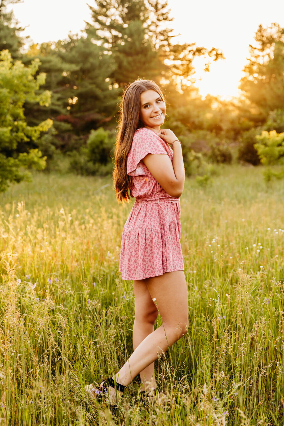 pretty teenage girl standing sideways with her knee popped and hand resting on her shoulder as she looks over when the sun sets behind her in a park near Oshkosh