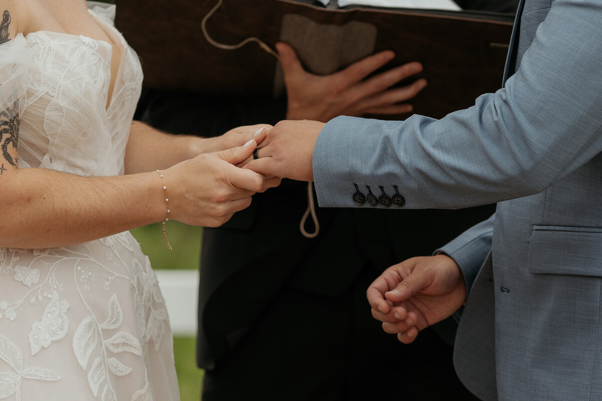 Close-up of bride and groom exchanging wedding rings during their summer outdoor ceremony at the Barn at Blackstone National. The bride's lace gown and delicate bracelet, along with the groom's light blue suit, are prominently featured.