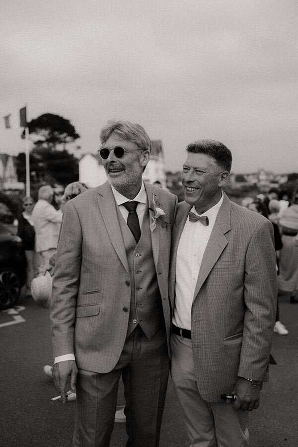 Patrick et un invité souriant. Un moment complice lors du mariage, capturé en noir et blanc.