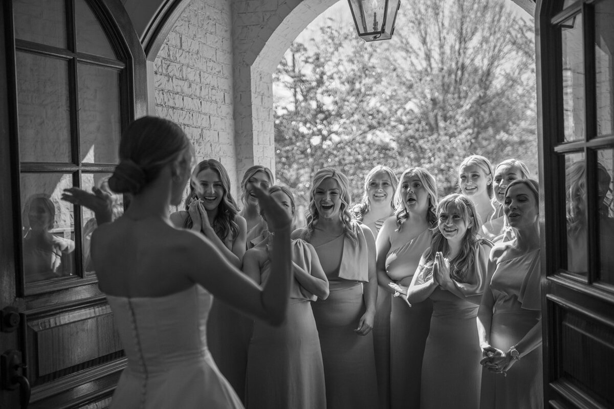 Black and white photo of the bride showing her wedding dress to her bridesmaids, who react with excitement and smiles during a first look at Burritt on the Mountain in Huntsville, Alabama, photographed by Perry Hancock.
