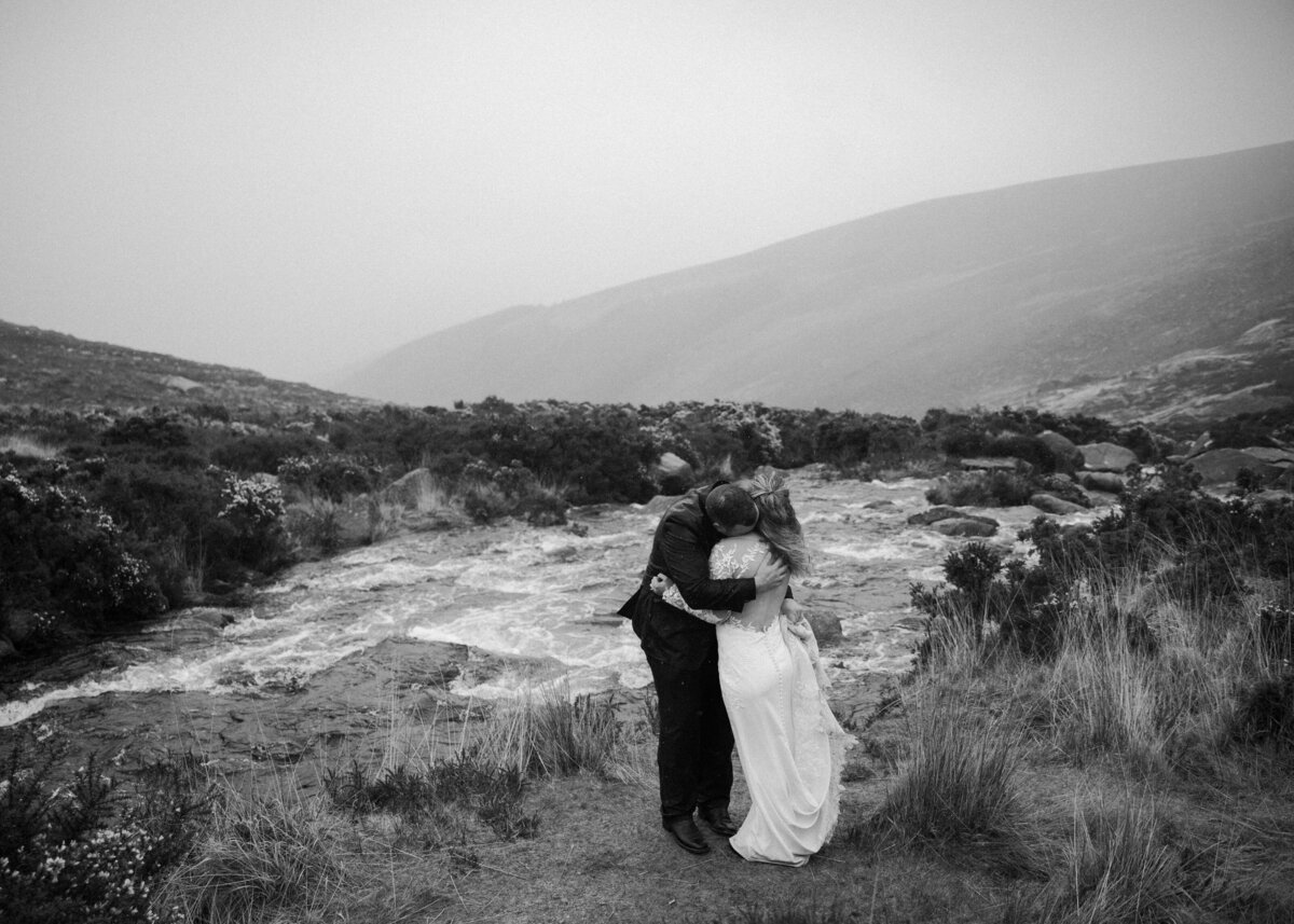 A black and white photo of a couple embracing in a windswept moorland landscape