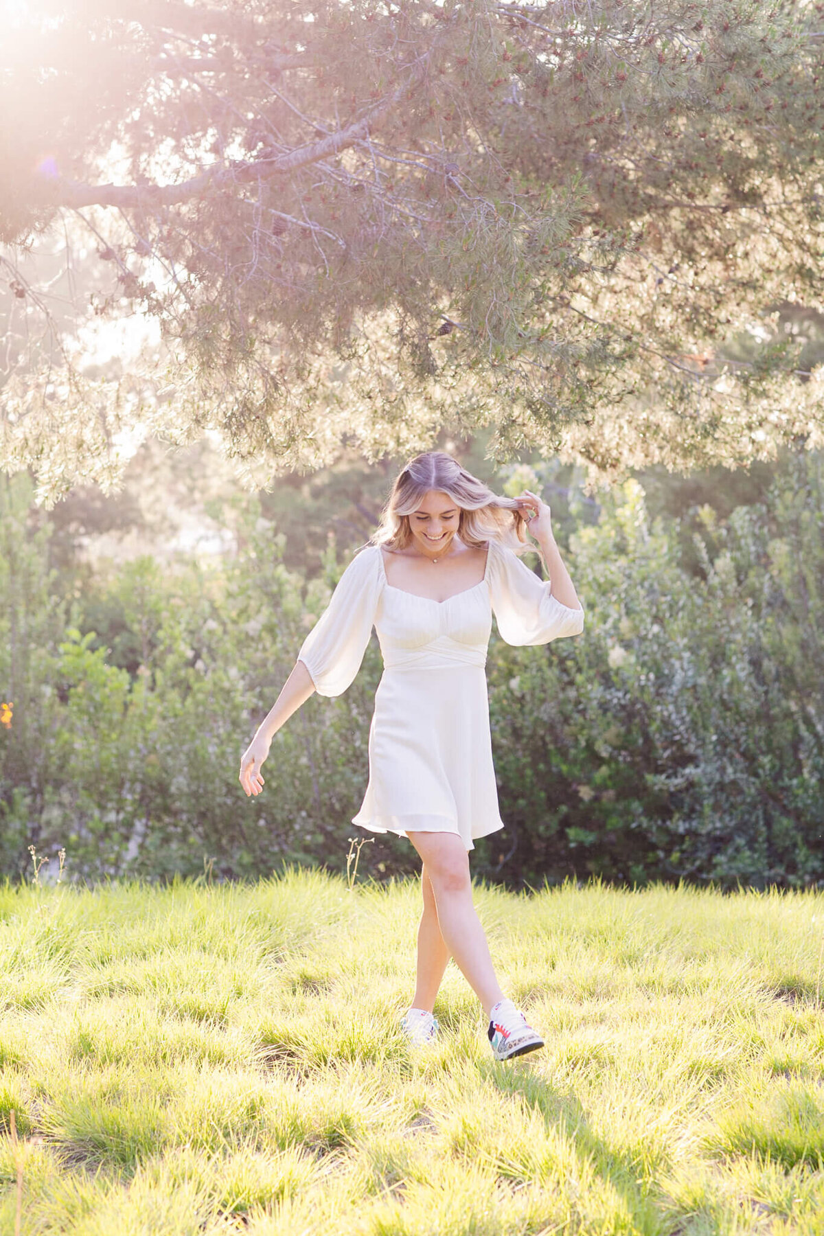 A woman in a white dress walks gracefully on sunlit grass, surrounded by lush greenery. Her hair is flowing in soft waves, and her expression is peaceful. Dappled sunlight filters through the trees above, casting a serene glow.
