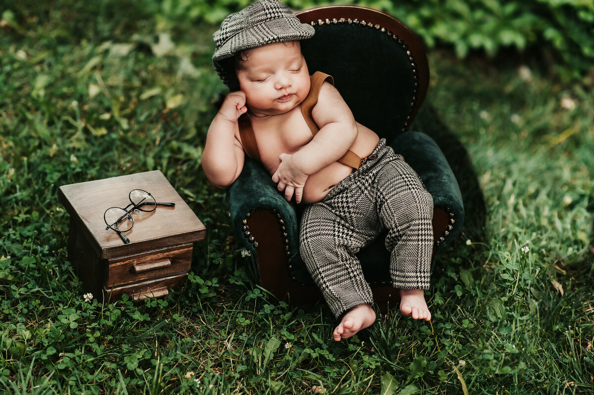 Infant boy sleeping at Greater Toronto newborn photo shoot outdoors on a green chair in a cute plaid hat and pants outfit.