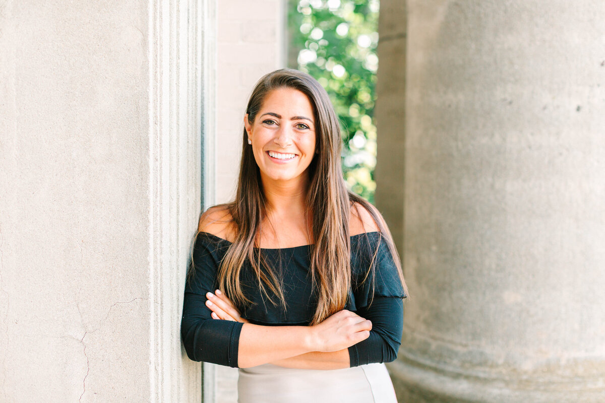 A woman with long brown hair and a smile leans against a stone column, arms crossed. She wears a black off-the-shoulder top and a light-colored skirt, with greenery visible in the background.