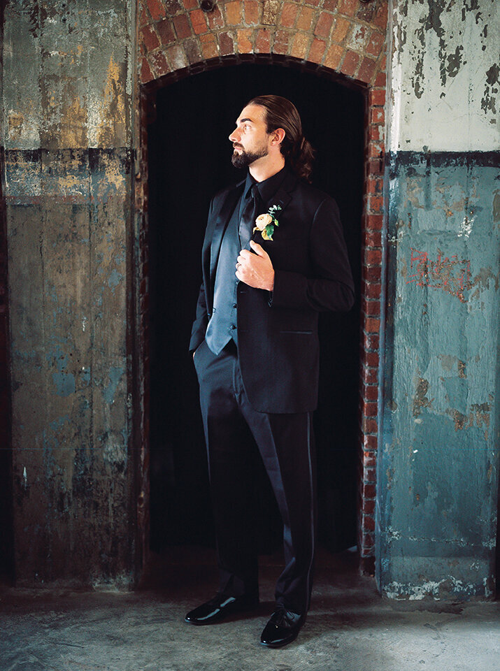 A groom wearing a black tuxedo stands in a doorway along a weathered brick and wooden wall