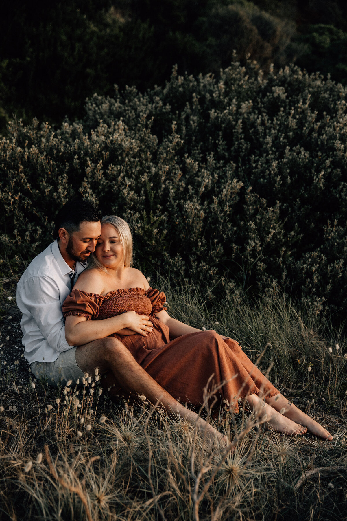 Expectant couple sit in beach grass and cuddle