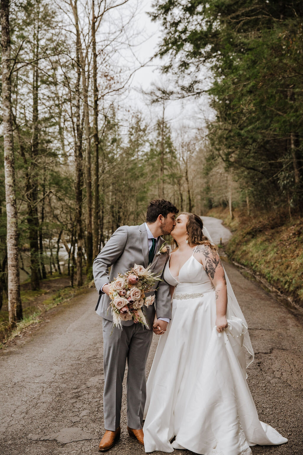 Bride and groom share a kiss on a forest-lined road in Greenbrier, holding a wedding bouquet during their Gatlinburg elopement.
