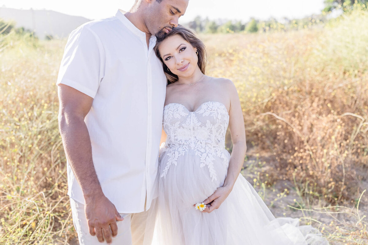 A couple stands in a sunny field. The woman, wearing a white lace dress, leans against the man, who is in a light shirt. They appear content and serene, surrounded by tall grass and a clear sky. The woman holds a small flower.