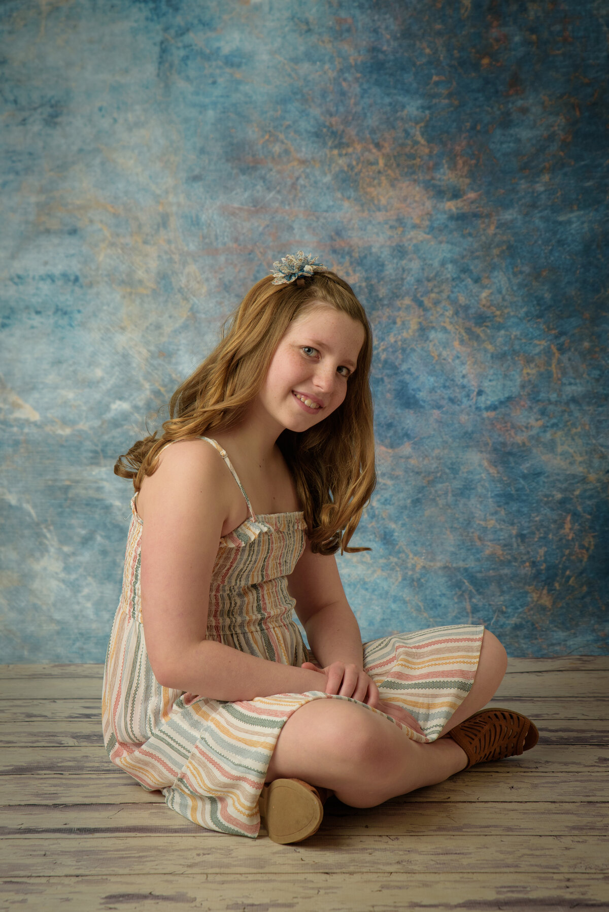 Beautiful young girl wearing a cream colored striped dress sitting on a wooden floor in front of a blue background at my home studio near Green Bay, Wisconsin