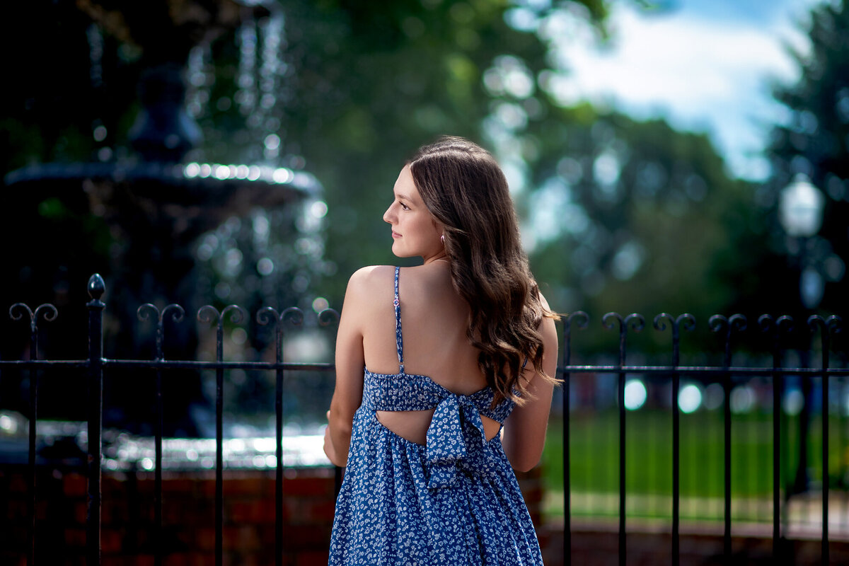 A woman in a blue floral dress leans on the iron fence of a fountain for a Pella Senior Photographer