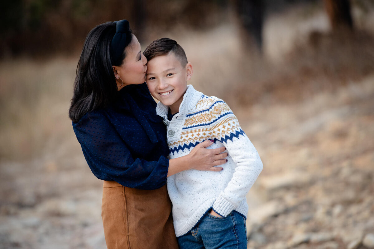 A mother kisses her smiling son in a white sweater