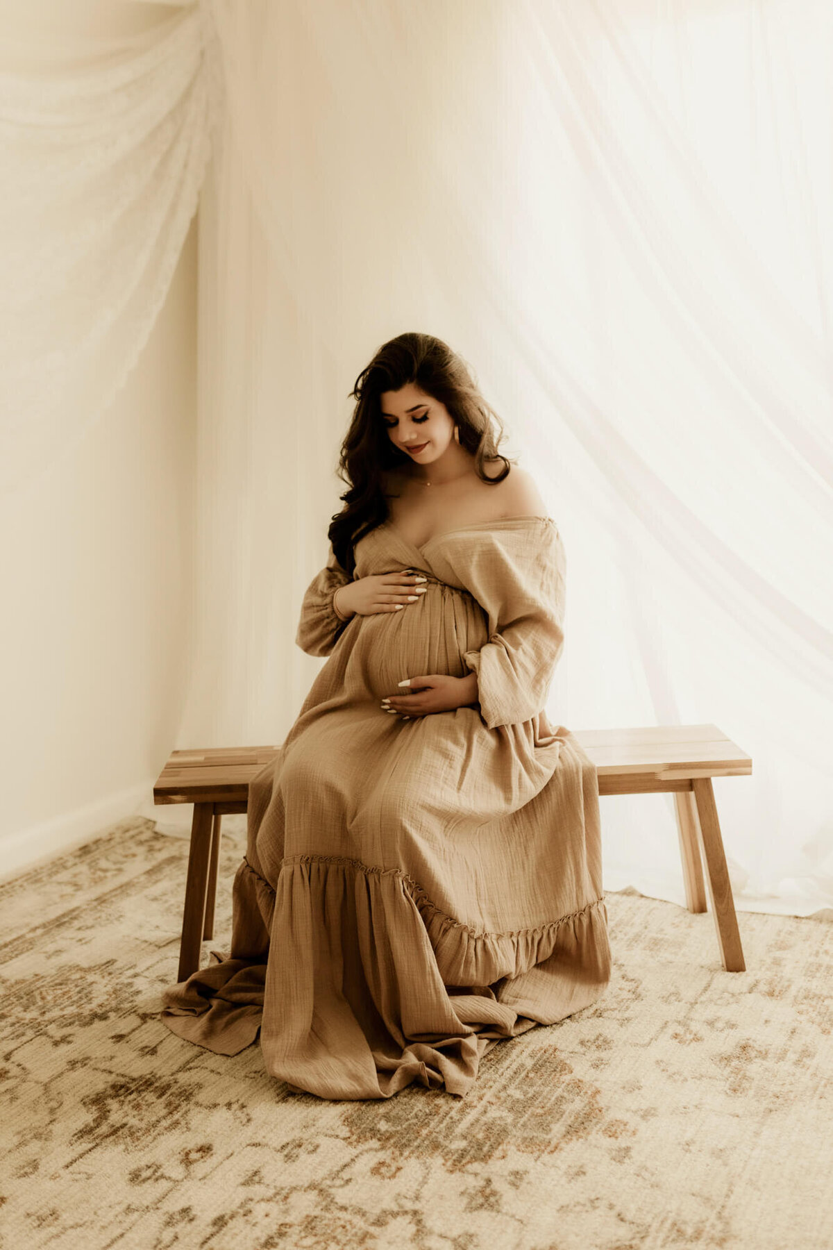 Expecting mother sitting on a bench while looking down at her  baby bump wearing an off-shoulder beige dress.