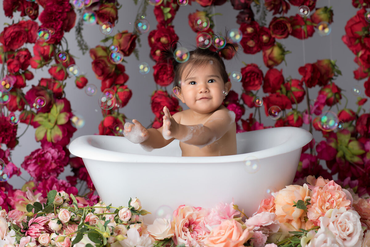 Surrounded by lush florals and floating bubbles, a baby plays in a white tub, clapping joyfully during a whimsical milestone session with a Dallas milestone photographer.