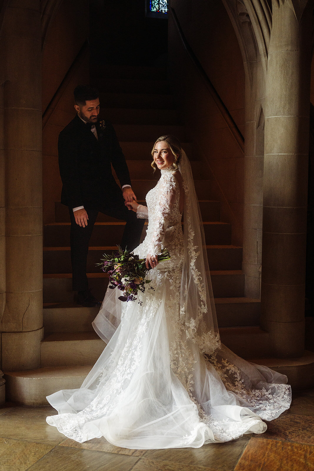 Bride in a long-sleeved lace gown descending a grand stone staircase, holding a dark bouquet, with the groom following behind.