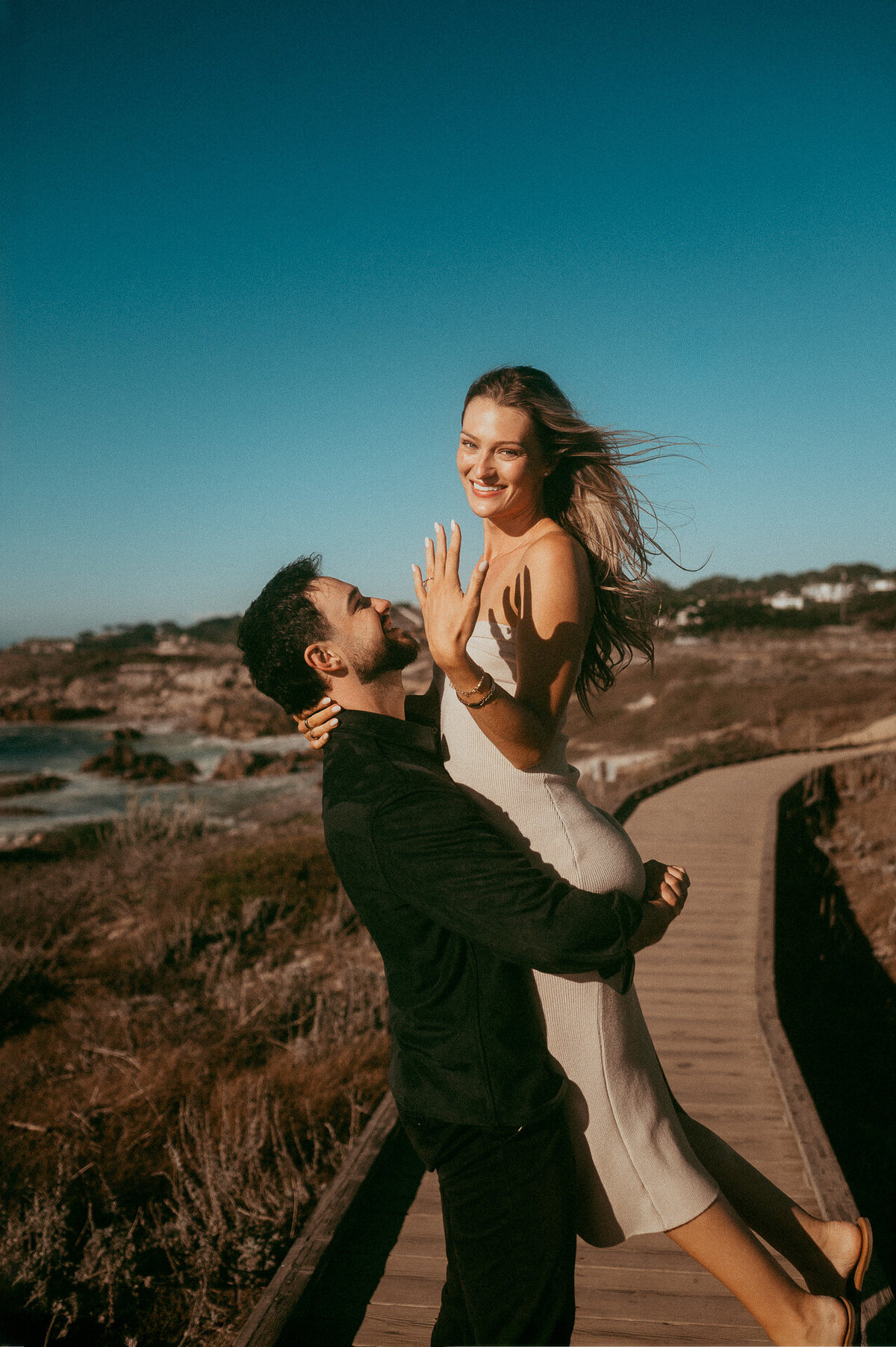 Fiancé lifting his fiancée on the Asilomar boardwalk in Pacific Grove as she shows off her ring