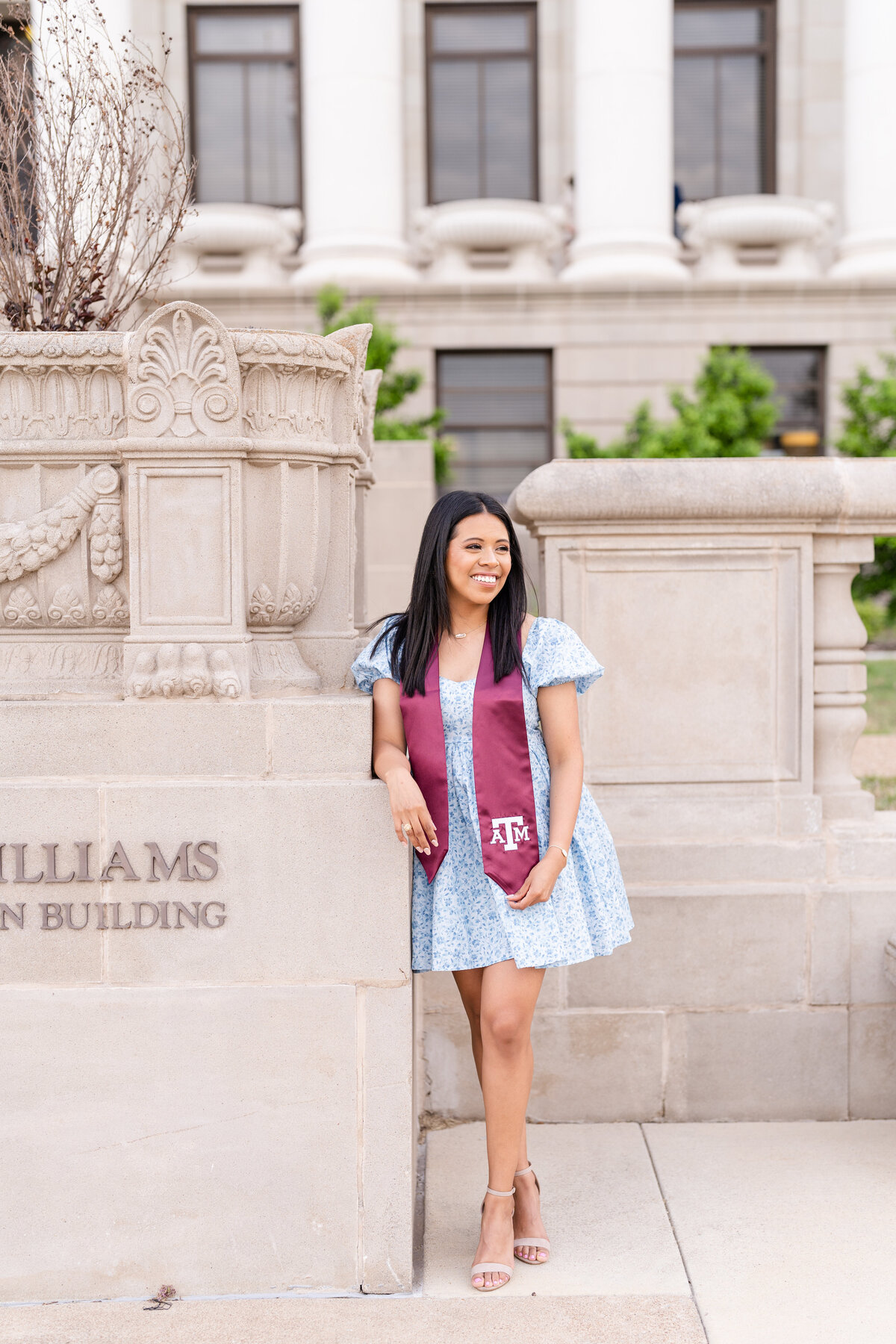 Texas A&M senior girl laughing away and leaning in front of Administration Building while wearing blue dress at maroon stole