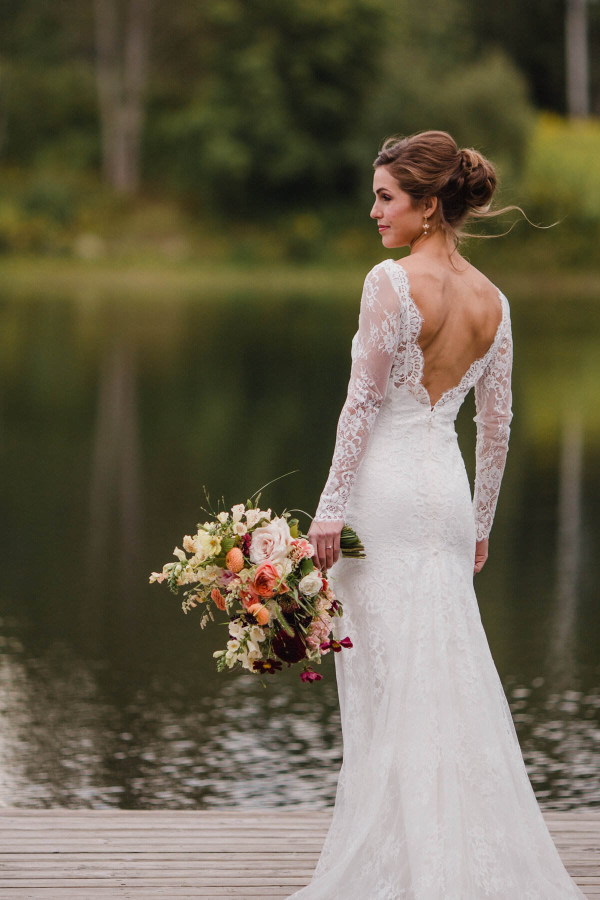 A bride in a lace dress stands by a lake holding a large flower bouquet.