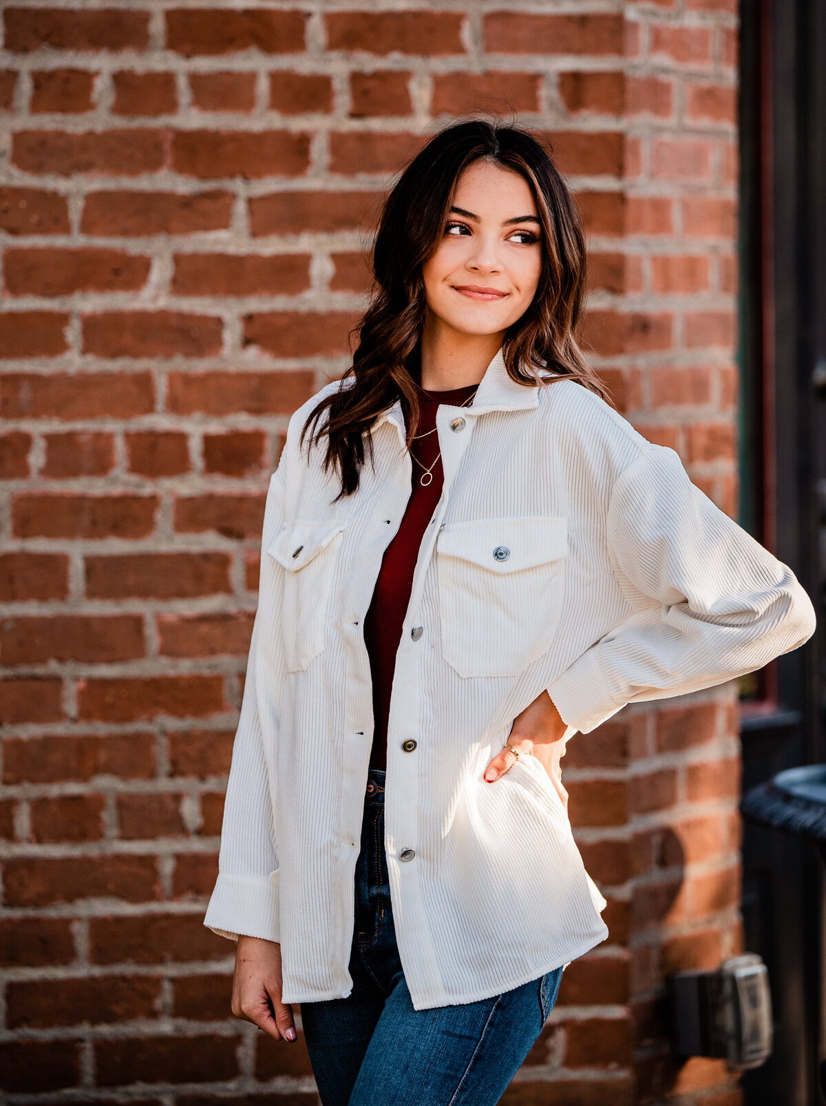 A senior wearing a white jacket stands in front of a red brick wall with her hand on her hip.