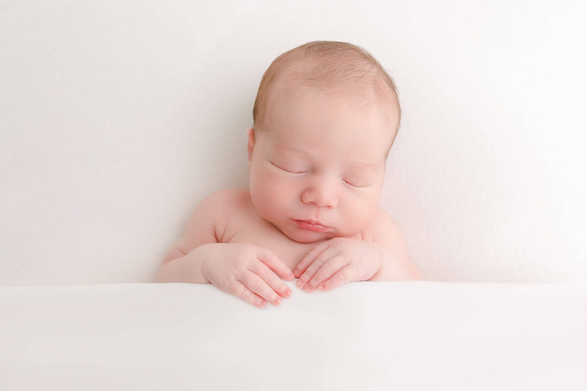 Newborn baby sleeping during newborn photo session tucked under a white sheet with his hands coming up over the sheet. Newborn photo session at Ashlie Behm Photography studio in Portland, Oregon.