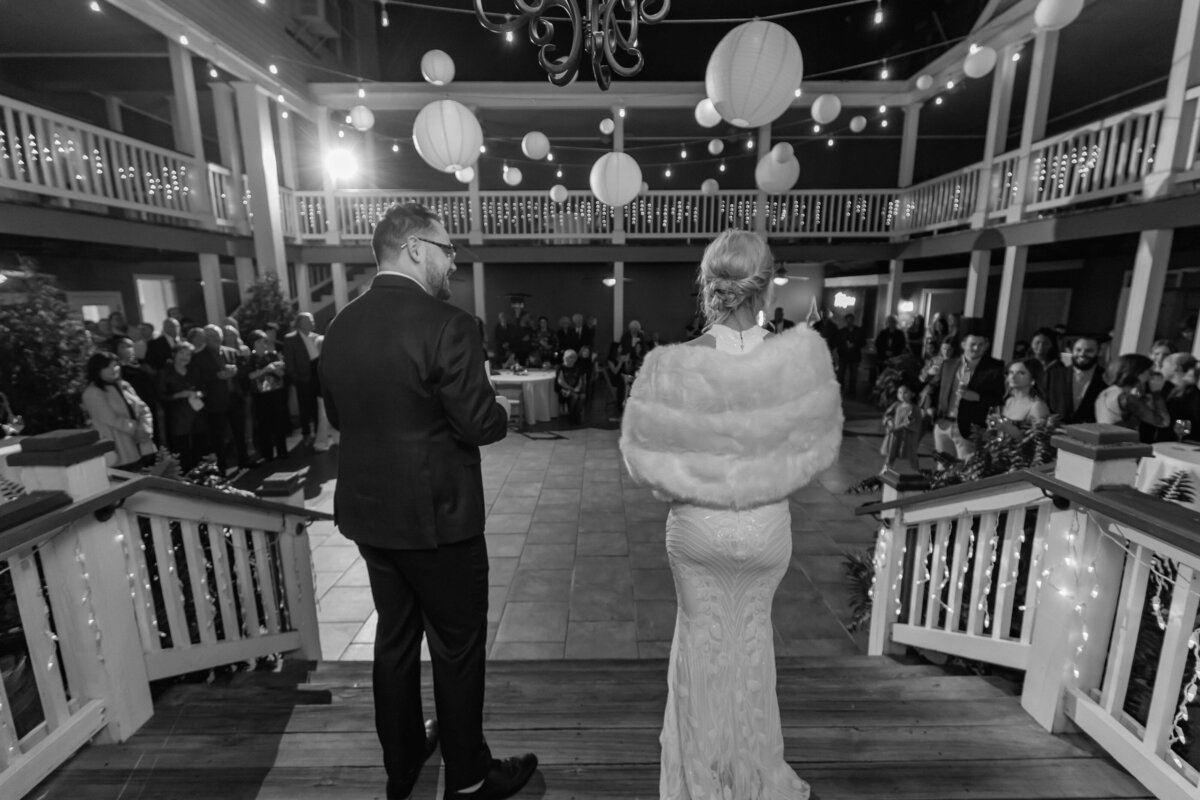 A bride and groom stand on a decorated staircase, facing a large crowd at a nighttime wedding reception. The venue has string lights and paper lanterns overhead, with guests gathered around the courtyard.