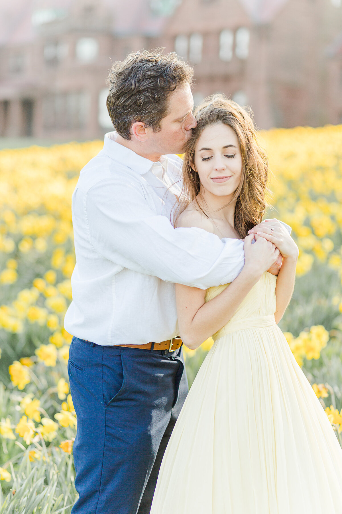 A woman stands with her back to her fiance amongst the daffodils at Salve Regina's Cliff Walk. The man has his arms wrapped around the woman's chest and is giving her a kiss atop of her head. Captured by best Rhode Island Wedding Photographer Lia Rose Weddings