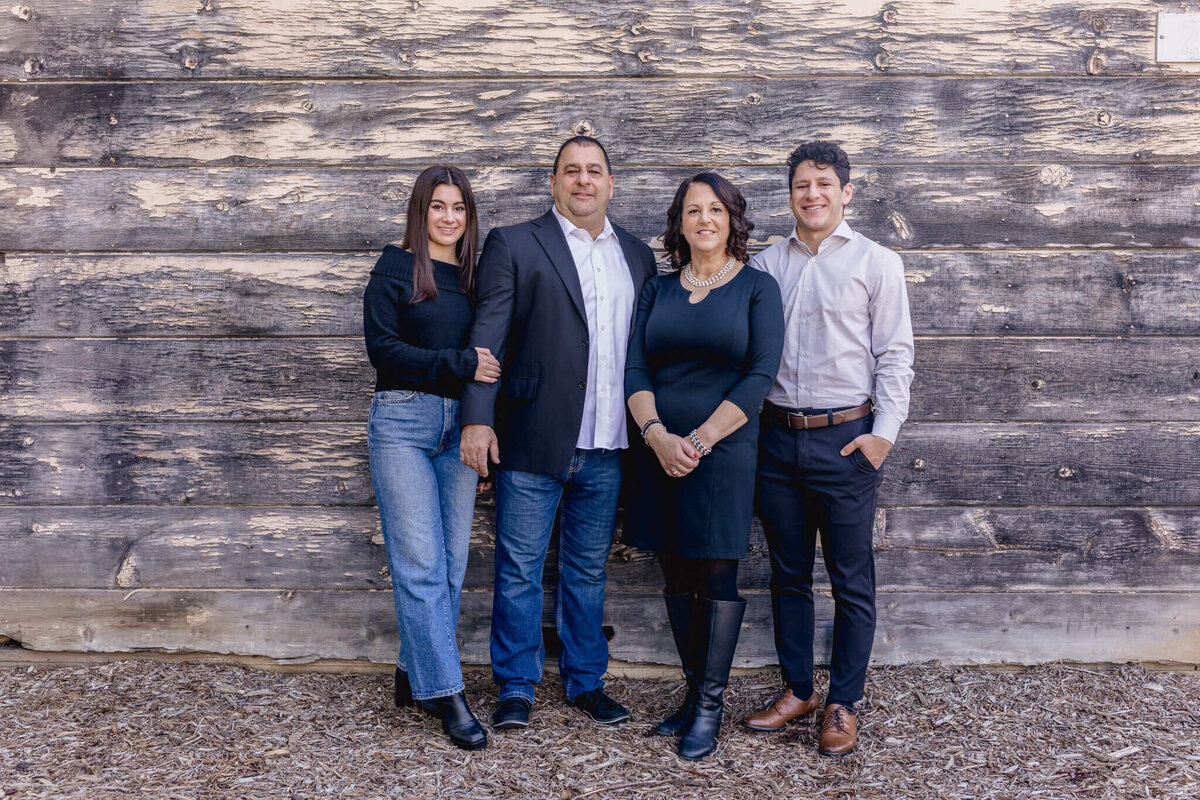 A family of four with dark hair standing close to each other in front of a long, wooden wall, looking at the camera.