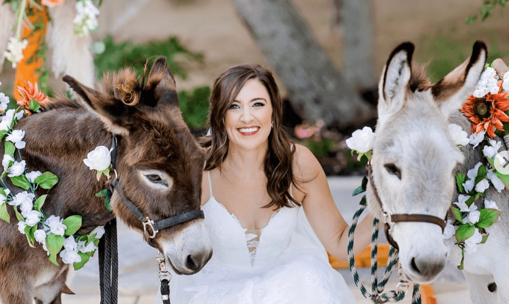 Bride with professional wedding makeup and curled hair poses with flower-adorned donkeys after bridal hair styling and manicure in San Antonio