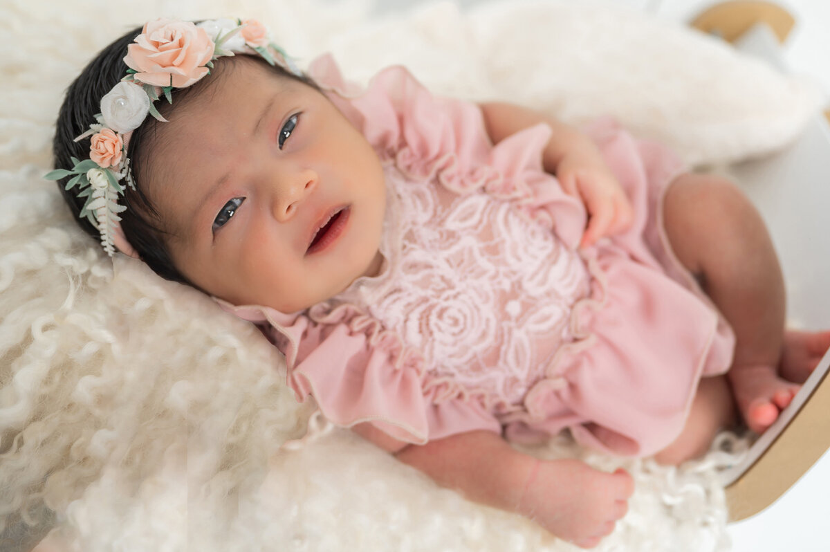 Baby with open eyes wearing a lacy soft pink dress and matching flower crown, lying on an off-white flokati rug.