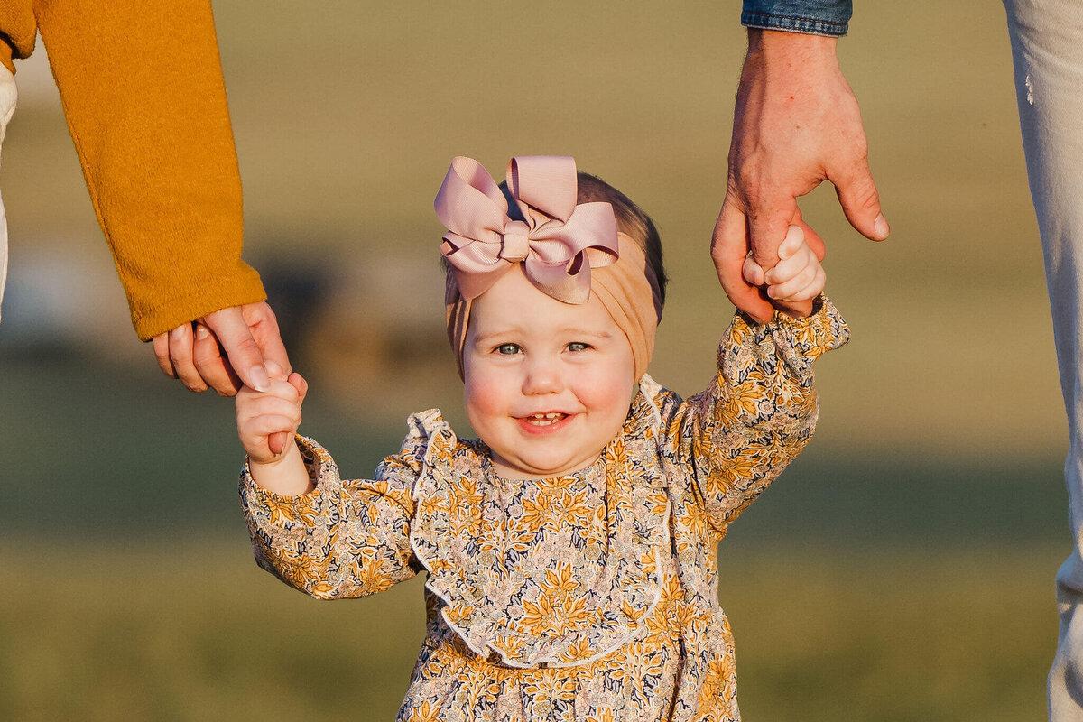 one year old girl holding parnets hands while waling through field at golden hour