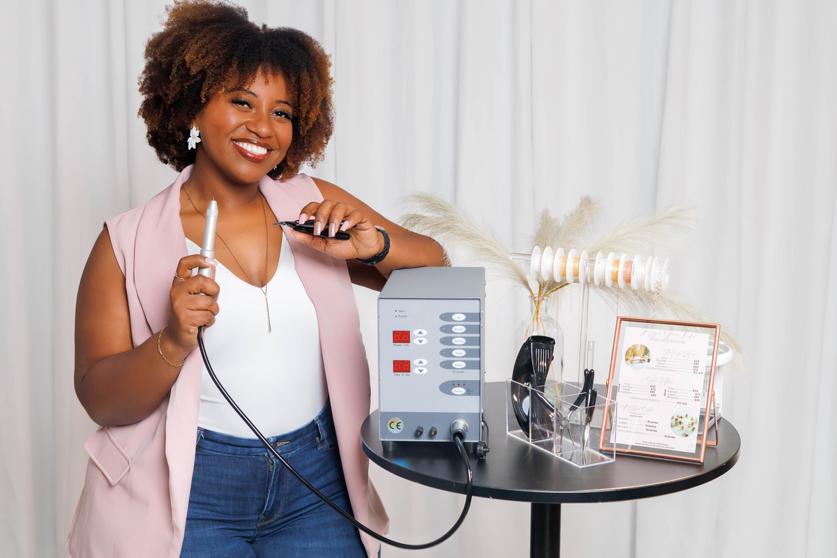 Jazzy smiling in front of the permanent jewelry welding machine at Jazzy's Glam Bar in Tampa, FL.