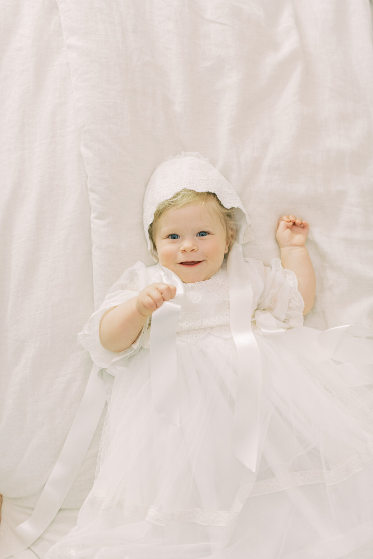 Joyful baby girl in heirloom white romper, playing with her baptism bonnet in a light-filled Oakville bedroom.