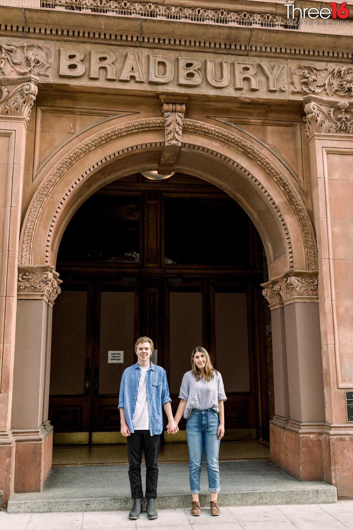 Engaged couple stand in front of the Bradbury Building holding hands