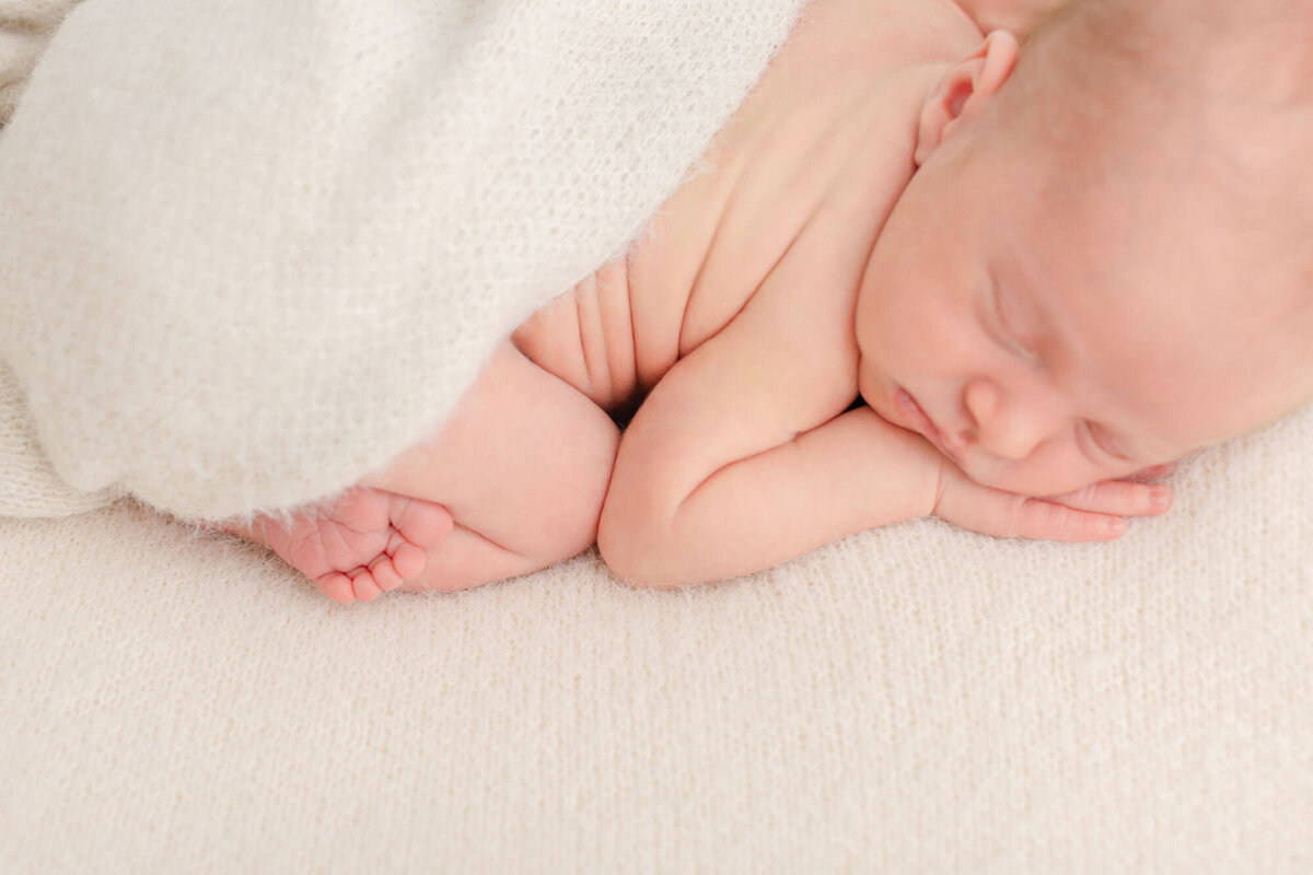 Image shot from above of a sleeping baby laying on his belly with a hand under his chin and his elbow and knee are touching creating some cute wrinkles on his back. He has a cozy beige blanket draped over his back.