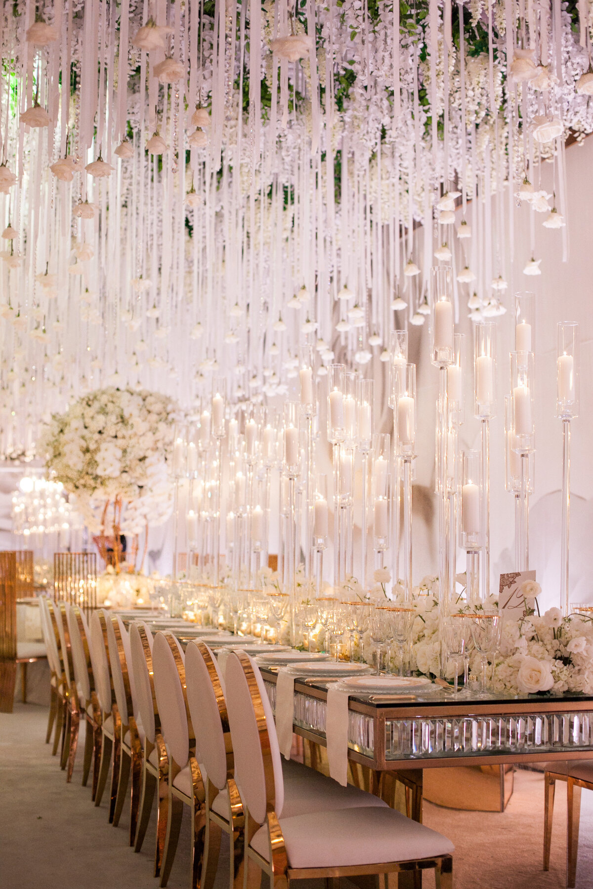 A lavishly decorated wedding reception features a long dining table adorned with numerous lit candles and elegant white floral arrangements. Gold-accented chairs line the table, and white flowers hang from the ceiling, creating a luxurious and romantic atmosphere.
