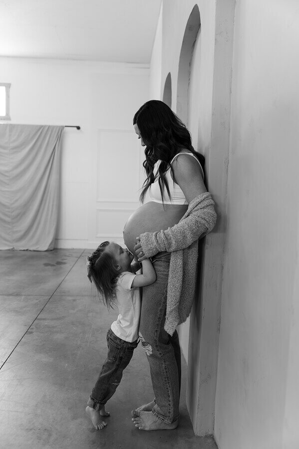 A pregnant mom holds the hands of her young child as she kisses her mom's belly.