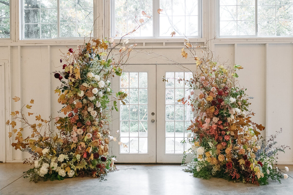 Large fall garden–inspired wedding ceremony floral installation featuring abundant roses, chrysanthemums, autumn foliage, and natural branches in warm tones of gold, burgundy, blush, and ivory. Arranged on either side of a set of French doors inside a bright white venue, creating a lush, organic, asymmetrical backdrop for an indoor ceremony.
