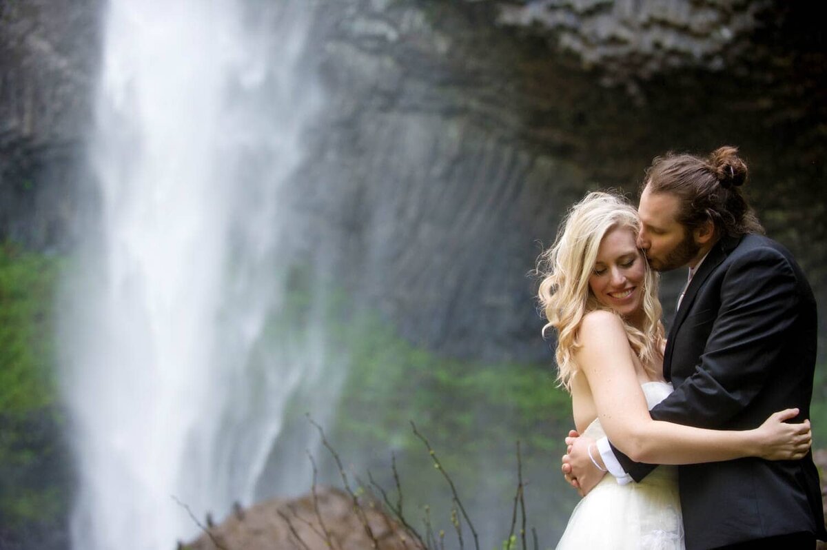 a man kisses a brides forehead with latourell falls in the background